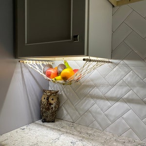 May include: A white hammock-style fruit basket hanging from a cabinet, filled with apples, oranges, avocados, and bananas. A brown owl figurine sits on a gray countertop below the basket.