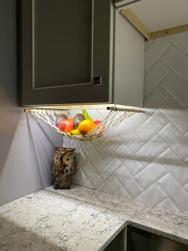 May include: A white hammock-style fruit basket hanging from a cabinet, filled with apples, oranges, avocados, and bananas. A brown owl figurine sits on a gray countertop below the basket.