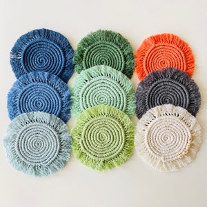 May include: A collection of nine handmade macrame coasters in various colors, including blue, green, and orange. Each coaster features a spiral design with a fringed edge. One coaster has wooden beads with the letters "M, Y, R, A".