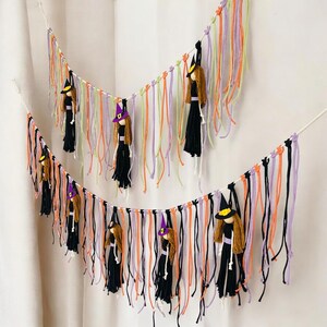 May include: Two Halloween witch garlands. Each garland features small witch dolls hanging from colorful yarn strands in orange, purple, black, and green. The witches wear black dresses and pointed hats with yellow or purple accents.