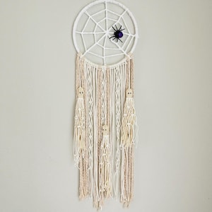 Peut inclure: Un attrape-rêves en macramé blanc avec un motif de toile d'araignée et une araignée violette. L'attrape-rêves présente des glands crème et beiges avec des perles en bois en forme de fantômes, parfait pour Halloween.
