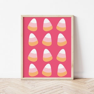 May include: A framed print with a repeating pattern of candy corn sweets on a vibrant pink background. Each candy corn is white, pale orange, and yellow. The print is in a light wood frame and is displayed on a white wall.