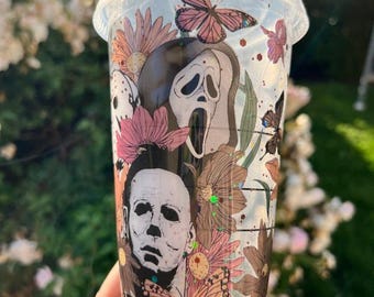 May include: Clear plastic tumbler with a lid and green straw. The tumbler features a design with images of Michael Myers and the Ghostface mask, surrounded by flowers and butterflies in shades of pink, brown, and beige.