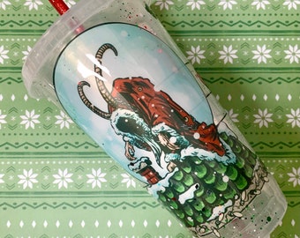 May include: Clear plastic tumbler with a red straw and a lid. The tumbler features a cartoon illustration of a creature in a Santa Claus outfit. The design includes a red coat, white beard, and green scales. The cup is decorated with glitter.