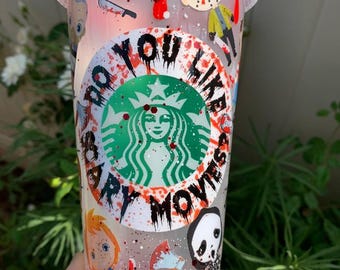 May include: A clear plastic tumbler with a red lid and a straw. The cup is decorated with horror movie characters, blood splatter, and the text "Do You Like Scary Movies?" around the Starbucks logo. The bottom of the cup has red glitter.