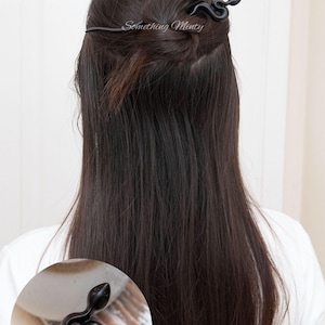 May include: Black snake hair stick with a decorative snake head. The stick is being used to hold a woman's hair in a bun.