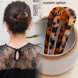 May include: Two brown and black tortoise shell hair clips with gold lettering that says "AA". The clips are in a white ceramic bowl.