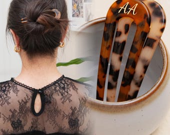 May include: Two brown and black tortoise shell hair clips with gold lettering that says "AA". The clips are in a white ceramic bowl.