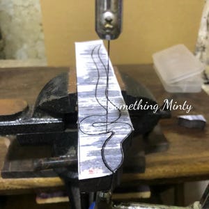 May include: A close-up of a scroll saw cutting a snake design out of a piece of metal. The metal is clamped in a vise and the saw is cutting through the design. The text "Something Minty" is visible in the background.