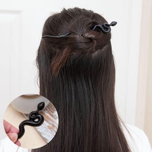 May include: Black snake hair clip with a long, thin, black metal pin. The clip is in the shape of a snake with a curled tail. The clip is being used to hold a woman's hair in a bun.