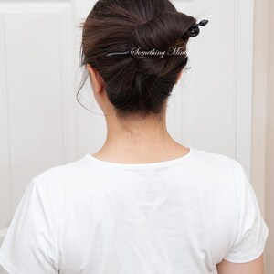 May include: A woman with dark brown hair styled in a bun with a black hair stick. The hair stick has a decorative design at the end. The woman is wearing a white t-shirt.