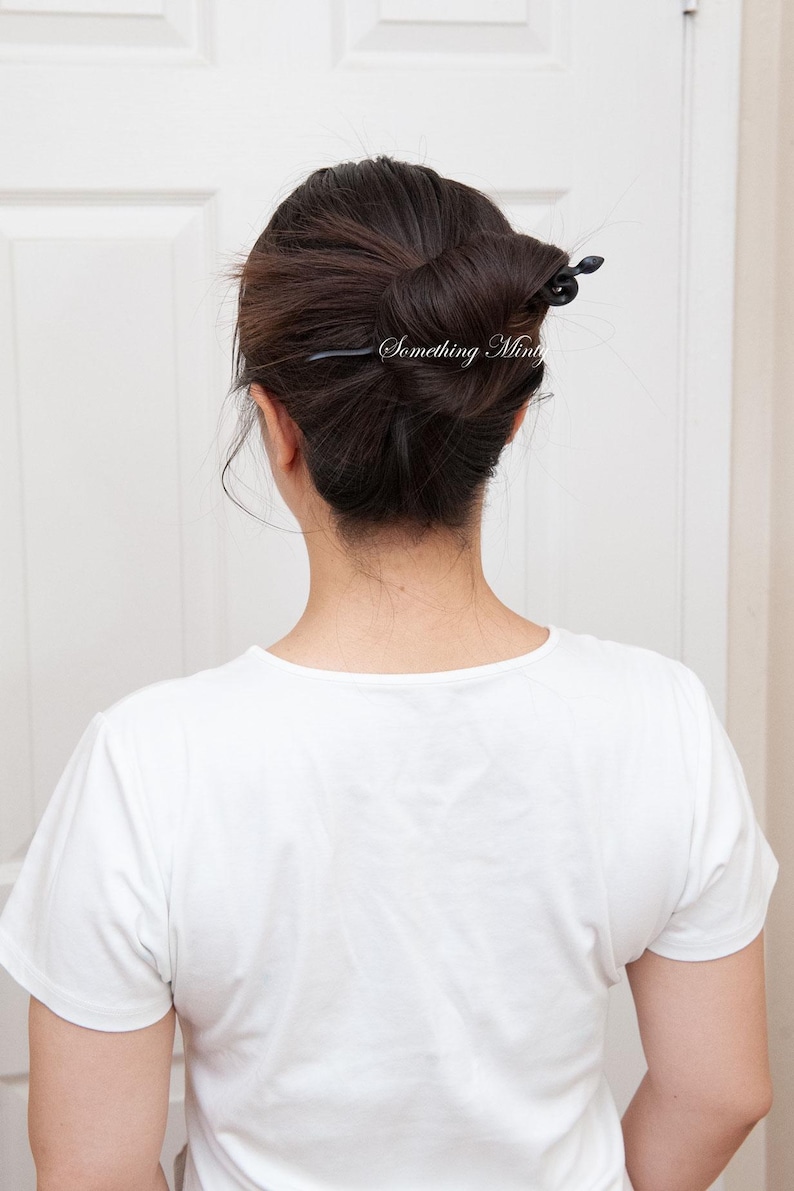May include: A woman with dark brown hair styled in a bun with a black hair stick. The hair stick has a decorative design at the end. The woman is wearing a white t-shirt.