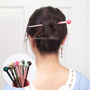 May include: A woman with dark brown hair is wearing a light blue shirt with white lace trim. She has a pink hair stick with a round pink bead on the end in her hair. The hair stick is in a bun. The text "Something Minty" is visible in the image.