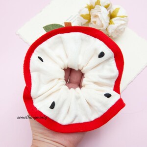 May include: A white and red apple-shaped scrunchie with black seeds. The scrunchie is made of soft fabric and is ideal for holding hair.