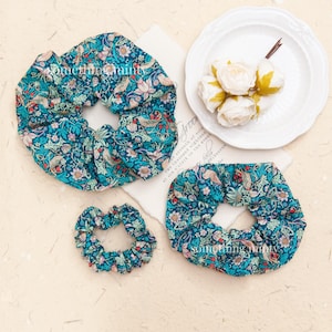 May include: Three teal blue fabric scrunchies with a floral pattern. The scrunchies are arranged on a white surface with a piece of paper with text.