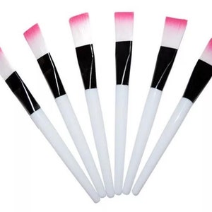 May include: Set of six white makeup brushes with black handles and pink bristles. The brushes are arranged in a fan shape.