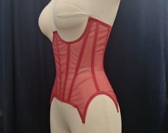 May include: A red corset displayed on a mannequin. The corset is made of sheer fabric with vertical boning and a scalloped hemline. The garment is designed to cinch the waist and create an hourglass silhouette. The corset is a vibrant shade of red.