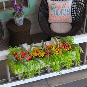May include: A miniature porch scene featuring two black lattice chairs, a small brown side table with a purple floral arrangement, and a white planter box filled with green, orange, and yellow faux plants. A white pillow reads it's CUDDLE SEASON.
