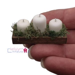 May include: Three white pillar candles arranged in a wooden tray with green moss.