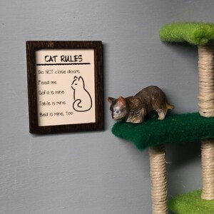 May include: A brown wooden framed sign with the text "CAT RULES" and a list of rules for cats, including "Do NOT close doors", "Feed me", "Sofa is mine", "Table is mine", and "Bed is mine, too". A small brown and white cat figurine is on a green cat tree.