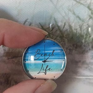 May include: A round, silver-toned pendant with a beach scene design. The image features a blue sky, turquoise water, and a sandy beach. The words "Beach Life" are written in a script font across the image.