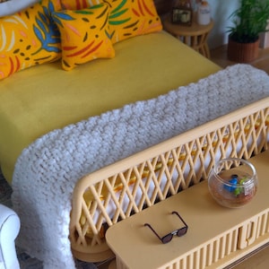 May include: Miniature bedroom scene with a yellow bedspread and pillows with colorful leaf patterns. A white knitted blanket is draped over the bed. A small side table holds a fishbowl and sunglasses. The bed frame and side table are made of light brown wicker.