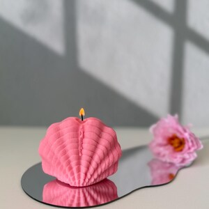 May include: A pink shell-shaped candle with a lit wick, sitting on a silver, wavy, reflective surface. The candle is on a white surface with a pink flower in the background.