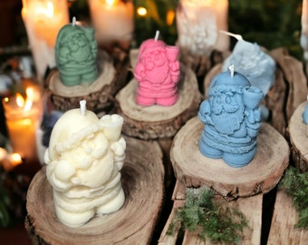 May include: A collection of Santa Claus-shaped candles in various colors, including cream, pink, blue, and green. The candles are displayed on rustic wooden slices, with lit candles and bokeh lights in the background, creating a festive holiday atmosphere.