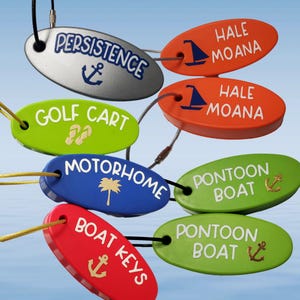 May include: A collection of oval-shaped floating keychains in various colors including silver, orange, lime green, blue, and red. Each keychain features white text and nautical or travel-themed icons such as anchors, sailboats, palm trees, and flip-flops.