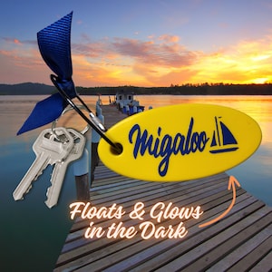 Puede incluir: Un llavero de forma ovalada amarillo con el texto "Migaloo" en letras azules y un pequeño gráfico de velero. El llavero está unido a un llavero plateado con dos llaves plateadas y una cinta azul. El texto "Floats & Glows in the Dark" está escrito debajo del llavero.