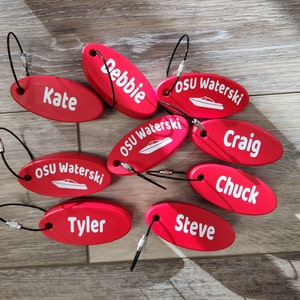 Può includere: Nove portachiavi ovali rossi con testo e grafica bianchi. I portachiavi presentano nomi come "Debbie", "Kate", "Craig", "Chuck", "Steve" e "Tyler". Tre portachiavi presentano il testo "OSU Waterski" con una piccola grafica di una barca.