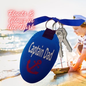 Pode incluir: Um porta-chaves azul "Captain Dad" com um desenho de âncora vermelha, preso a um porta-chaves prateado com chaves. O porta-chaves tem o texto "Floats & Glows in the Dark". Uma criança brinca com um veleiro de brinquedo no fundo.