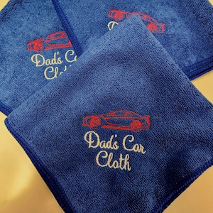 May include: Three square blue microfiber cloths featuring an embroidered red outline of a sports car above the text "Dad's Car Cloth" stitched in light yellow thread. The cloths have a textured surface and are stacked in an overlapping arrangement.