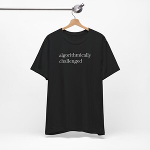 May include: A black short-sleeved t-shirt featuring the white text "algorithmically challenged" printed in a serif font on the chest. The garment is displayed on a wooden hanger against a plain background, showcasing a crew neckline.