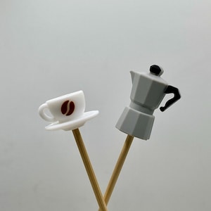 May include: A white miniature coffee cup with a brown coffee bean design and a gray miniature stovetop espresso maker, both attached to wooden skewers. The objects are set against a light gray background.