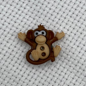 May include: A brown plastic monkey button with a light brown face and two black eyes. The monkey is sitting with its arms outstretched and its legs bent.