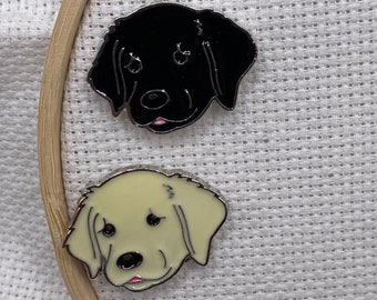 Puede incluir: Dos pines de esmalte con caras de perro. Uno es negro con contorno plateado, el otro crema con contorno plateado. Ambos pines tienen lenguas rosas, sobre un fondo de tela blanca.