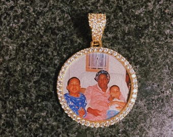 May include: A gold pendant featuring a round photo of three people. The pendant is encircled by small clear stones.
