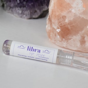 May include: A clear lip gloss tube with a white label that says "libra lip shine by starlightreads charming, artistic, peace loving." The tube is sitting on a white surface.