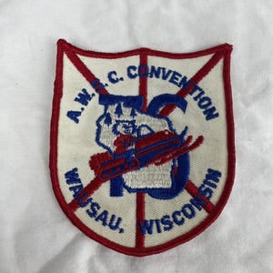 May include: A shield-shaped embroidered patch with a white background and red border. It features the text A.W.S.C. CONVENTION WAUSAU, WISCONSIN in blue, a large blue letter C, and a graphic of a small figure on a red snowmobile with pine trees.
