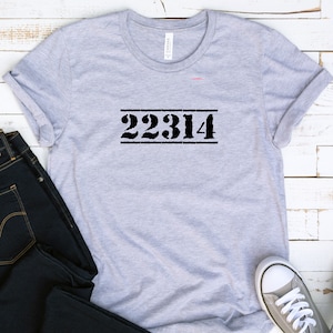May include: A light gray t-shirt with the black numbers 22314 printed in a stencil font. The numbers are centered and framed by two horizontal lines. The shirt is folded and displayed with dark blue jeans and a pair of gray sneakers.