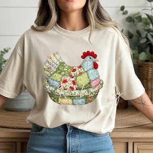 May include: Beige t-shirt featuring a patchwork chicken design. The chicken is composed of various floral and patterned fabrics, with a red comb and beak. The design is set in a patchwork basket, with the text "Rainbow Sky" below.