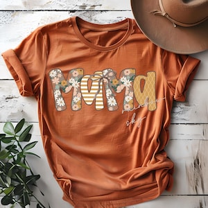 May include: An orange t-shirt featuring the word "MAMA" in a floral and striped design. The words "rainbow sky" are written in cursive below the word "MAMA". A brown hat is in the background.