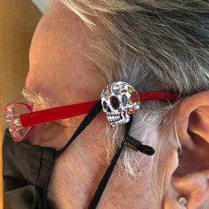 May include: A black face mask with a red and clear glasses frame. A black elastic strap with a colorful sugar skull charm is attached to the glasses frame.