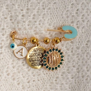 May include: A gold safety pin with charms, including a blue evil eye, a white charm with the letter A, a gold charm with Arabic script, and a gold charm with a green stone and the Arabic word for God.