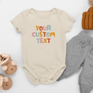 Peut inclure: Un body pour bébé à manches courtes de couleur crème avec le texte YOUR CUSTOM TEXT imprimé dans une police arrondie et colorée. Le body est présenté avec un pantalon de jogging gris, un bonnet en tricot marron et deux chaussons beiges.