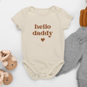 May include: A beige baby bodysuit with the text "hello daddy" in brown, and a brown heart. It is shown with grey jogging bottoms, a brown knitted hat, and brown baby booties.