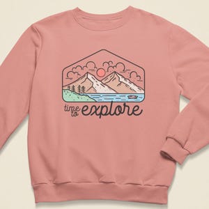 Peut inclure: Un sweat-shirt rose poudré avec un motif de montagne dans un hexagone. Le dessin comprend des montagnes, un lac, des arbres et un soleil, avec les mots "time to explore" en écriture noire.