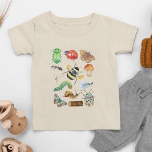 May include: A beige toddler t-shirt with a watercolour illustration of insects, including a green beetle, ladybug, butterfly, and bumblebee. The shirt is paired with grey sweatpants and a brown knitted hat.