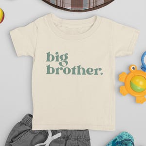 Peut inclure: Un t-shirt à manches courtes de couleur crème avec le texte "big brother" en lettres vertes.